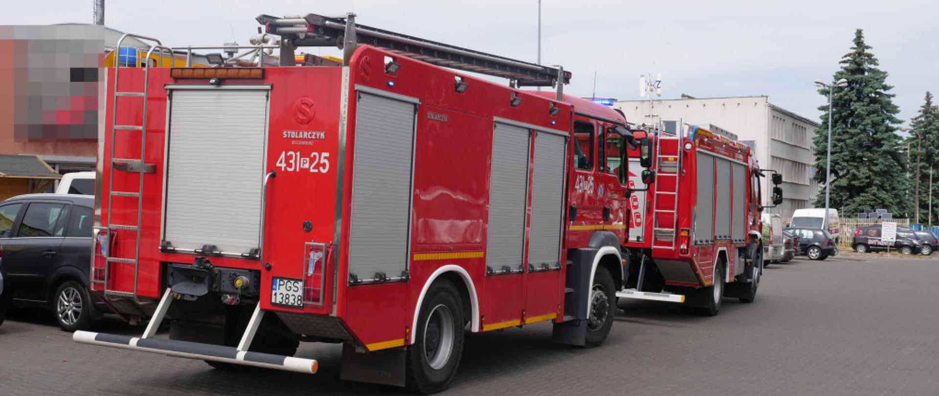 Zdjęcie przedstawia 2 wozy strażackie z włączonymi sygnałami błyskowymi. Samochody stoją na parkingu przed sklepem budowlanym.