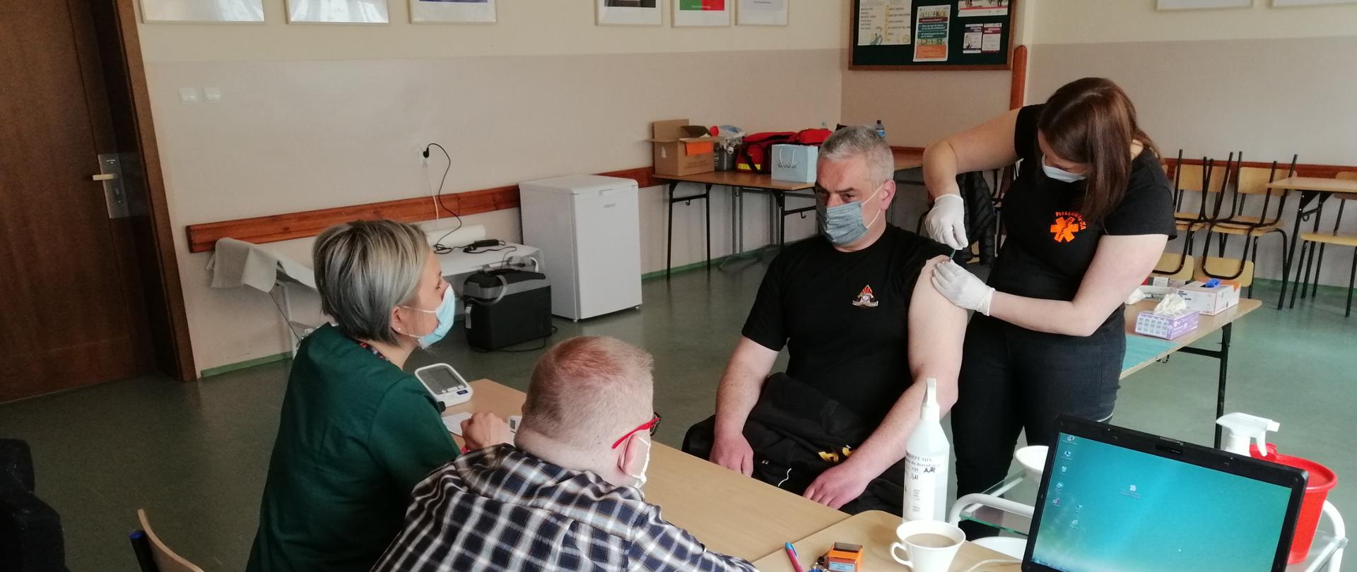 Na zdjęciu widzimy jak personel medyczny szczepi funkcjonariusza Państwowej Straży Pożarnej