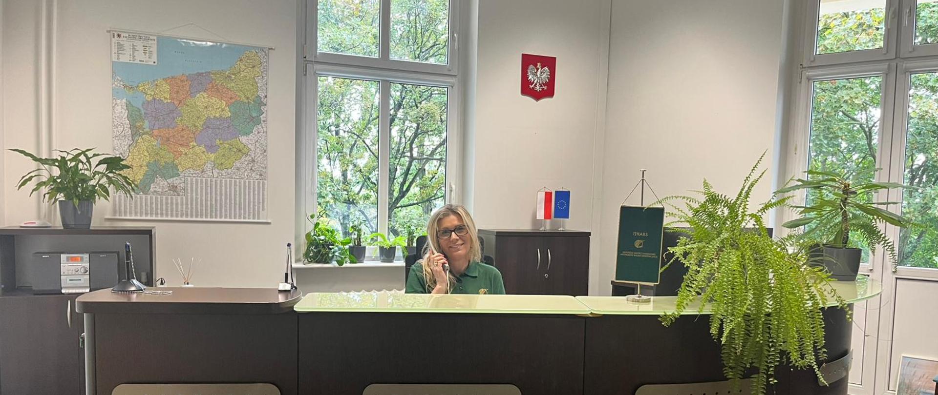 Pracownik służby cywilnej siedzi przy biurku, pracując przy dokumentach i komputerze, w tle widoczne godło państwowe.”