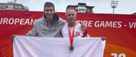 Dwaj mężczyźni reprezentujący Polskę stoją na podium sportowym na tle czerwonej ścianki z napisem European Police & Fire Games - Vigo 2026. Mężczyzna na stopniu nr 1 ma na sobie białą koszulkę z polskim godłem oraz medal na szyi, a mężczyzna po lewej (na stopniu nr 2) szarą bluzę z kapturem z grafiką dyscyplin sportowych. Wspólnie trzymają rozpostartą flagę Polski.