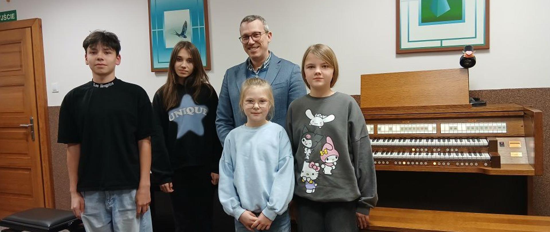 Zdjęcie przedstawia cztery młode osoby stojące obok dorosłego mężczyzny w jasnej koszuli i marynarce. Wszyscy pozują do zdjęcia w pomieszczeniu szkolnym. Po prawej stronie znajduje się konsola organów z widocznymi klawiaturami i pedałami. Osoby stoją w równym rzędzie, patrząc w stronę aparatu. W tle widać drewniane drzwi, krzyż na ścianie, zielony znak „Wyjście” oraz dwie zawieszone na ścianie ilustracje przedstawiające ptaki.