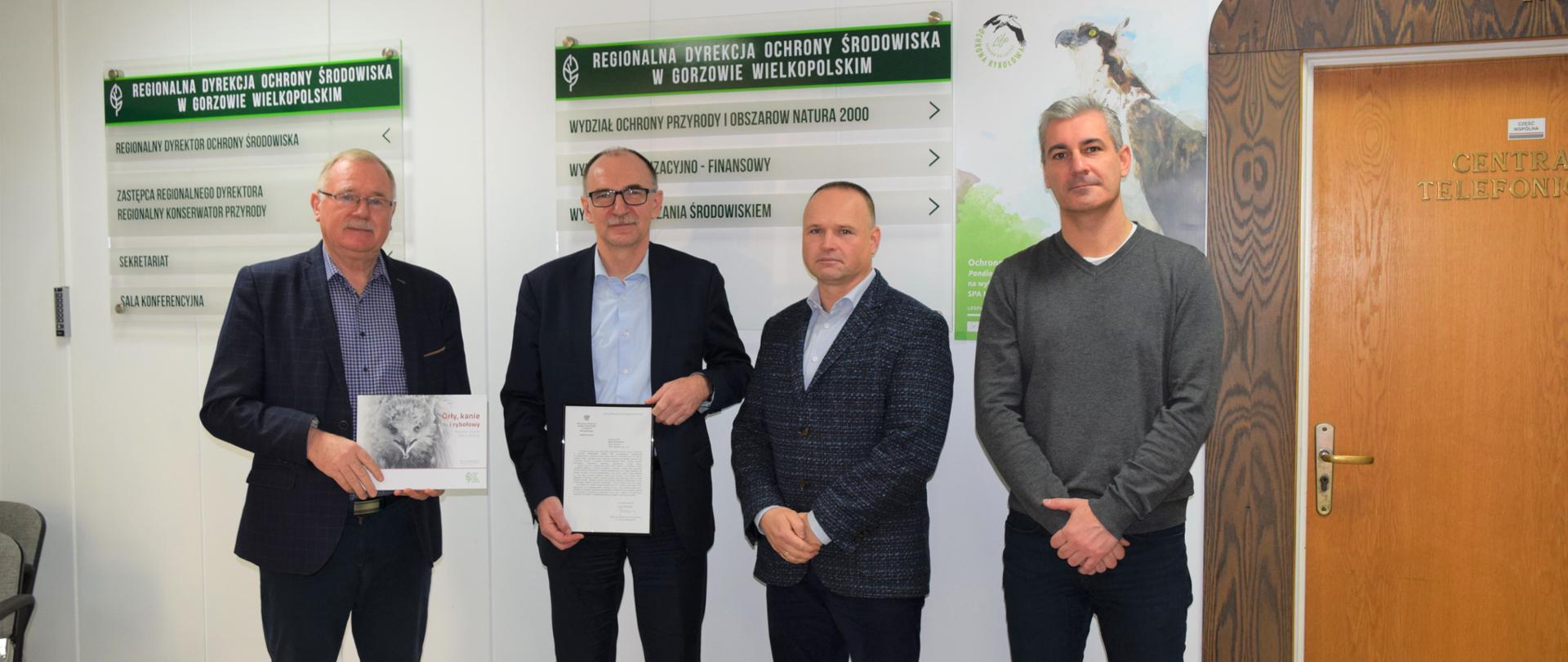 Czterech mężczyzn pozuje na tle ściany. Dwóch z nich trzyma w rękach książkę i ramkę z zapisaną kartką. Na ścianie widać napis Regionalna Dyrekcja Ochrony Środowiska w Gorzowie Wielkopolskim.
