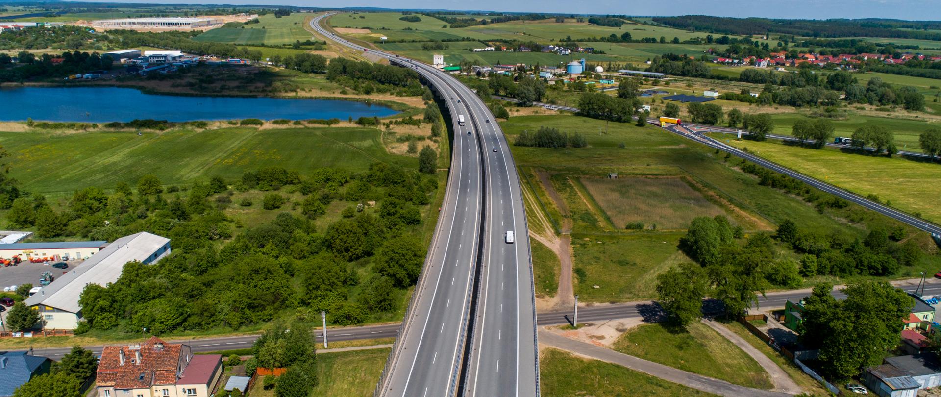 Droga dwujezdniowa na estakadzie poprowadzona nad drogą, linią kolejową i zbiornikiem wodnym. Widoczne zabudowania mieszkalne i przemysłowe oraz zieleń. Widok z lotu ptaka.