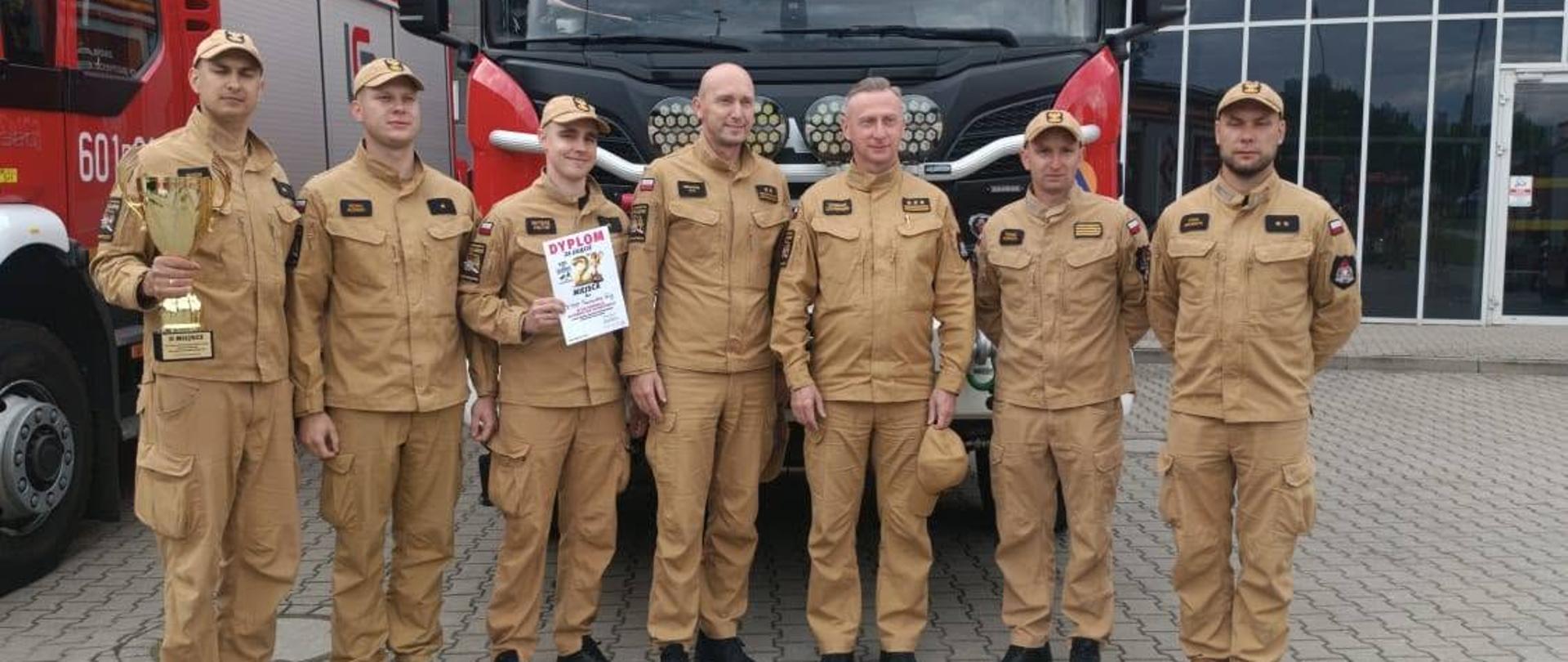 Oto szczegóły widoczne na fotografii:
Strażacy: Na pierwszym planie, przed wozem bojowym, stoi dumnie rząd siedmiu strażaków (prawdopodobnie reprezentacja danej jednostki PSP). Wszyscy są ubrani w jednolite, piaskowe mundury służbowe (koszule i spodnie) oraz czarne buty. Część z nich ma na głowach czapki z daszkiem w tym samym kolorze.
Nagrody: Strażacy świętują sukces. Mężczyzna po lewej stronie trzyma w dłoni duży, złoty puchar. Trzeci strażak od lewej trzyma z kolei biały dyplom, na którym wyraźnie widać napis „DYPLOM za zajęcie II MIEJSCA”.
Pojazdy ratownicze: Strażacy pozują bezpośrednio na tle przodu nowoczesnego, czerwonego samochodu pożarniczego (marki Scania z zabudową Szczęśniak, na co wskazuje napis na górze owiewki). Po lewej stronie kadru widać fragment kolejnego czerwonego wozu strażackiego z widocznym początkiem numeru operacyjnego (601...).
Tło i otoczenie: Grupa stoi na placu wyłożonym szarą kostką brukową. W tle znajduje się nowoczesny, duży budynek o charakterystycznej architekturze z licznymi przeszkleniami – konstrukcja dachu oraz elewacja przypominają nowoczesną halę sportową, widowiskową lub centrum szkoleniowe.