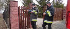 Dwóch strażaków OSP wrzuca ulotki informacyjne na temat szczepień przeciwko COVID-19 do skrzynki na listy. Posesja prywatna.
