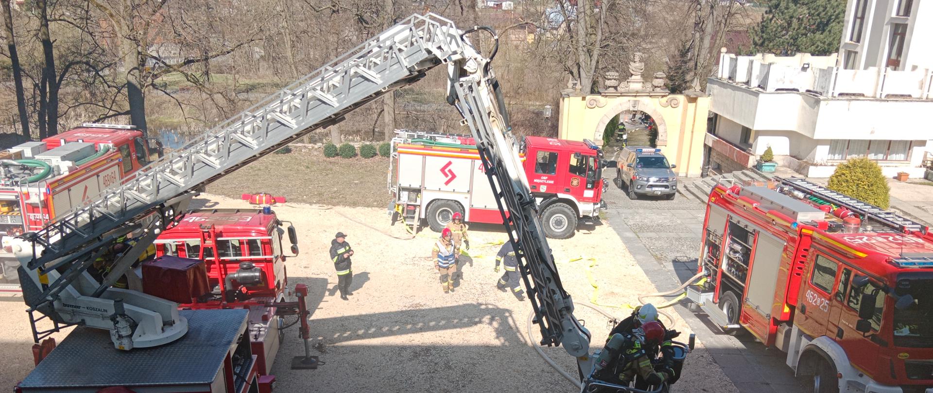 Widok z okna na duży plac. Na placu stoją trzy samochody ratowniczo-gaśnicze oraz podnośnik z koszem.