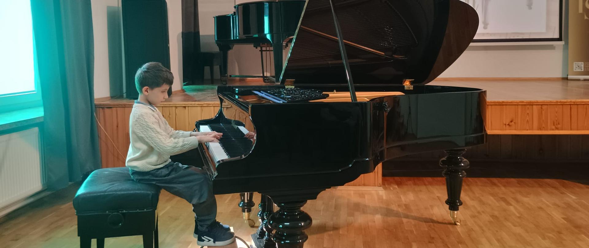 Zdjęcie przedstawiające wykonawców przy fortepianie w trakcie popisu uczniów klasy pani Małgorzaty Niewęgłowskiej, który odbył się w auli szkoły 28 maja 2025 roku.