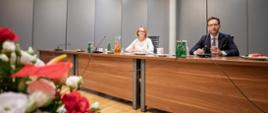 Minister Waldemar Buda oraz uczestniczka spotkania siedzą za stołem konferencyjnym. Na zdjęciu widoczny również bukiet kwiatów