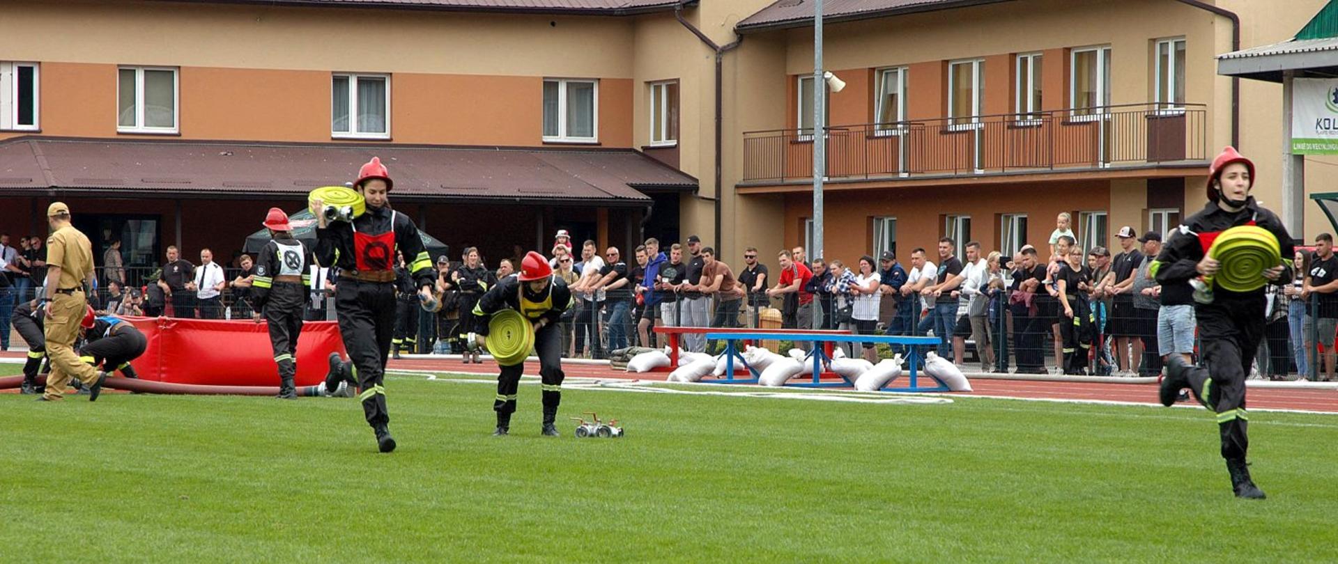 Zdjęcie zrobione na stadionie podczas zawodów sportowych drużyn Ochotniczych Straży Pożarnych. Trzy zawodniczki z kobiecej drużyny OSP biegnie po murawie stadionu, w rękach trzymają zwinięte węże pożarnicze. 