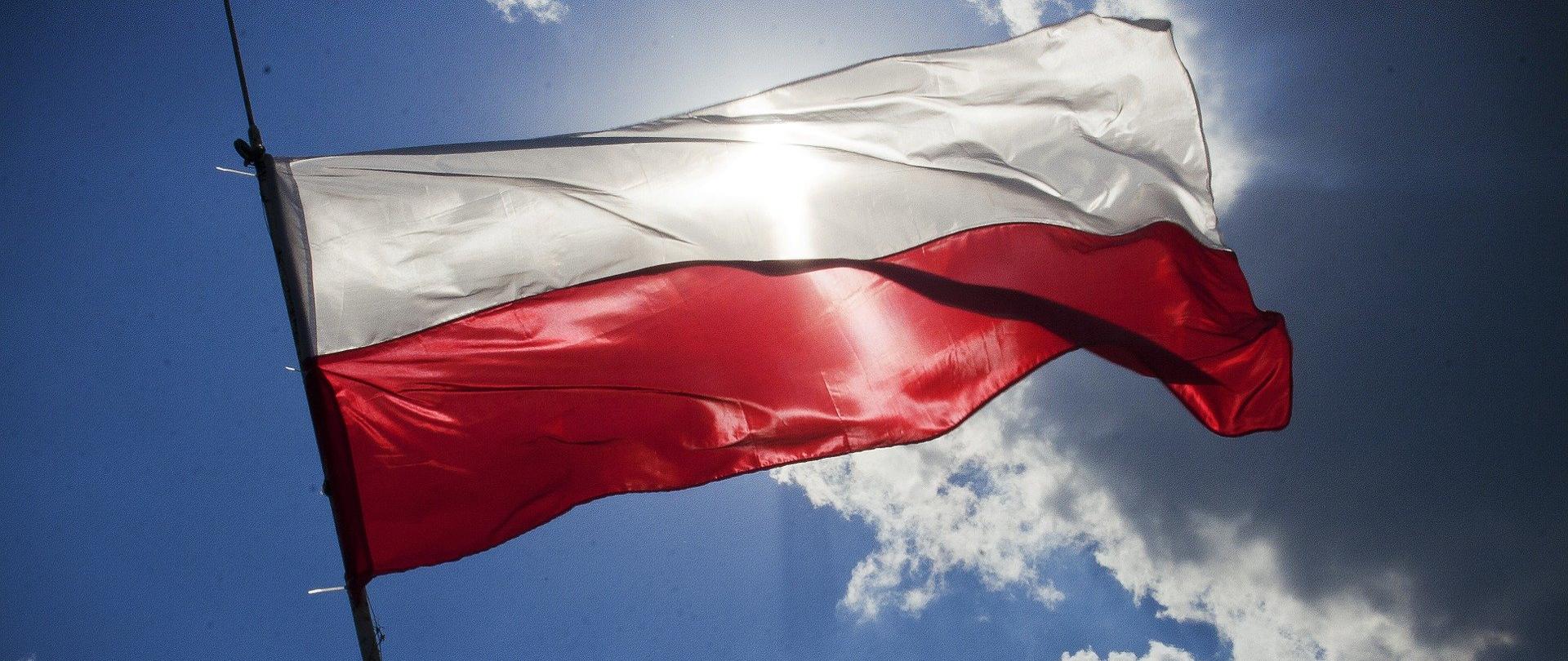 Zdjęcie przedstawia dużą, powiewającą na wietrze biało-czerwoną flagę RP. W tle błękitne niebo oraz chmury.