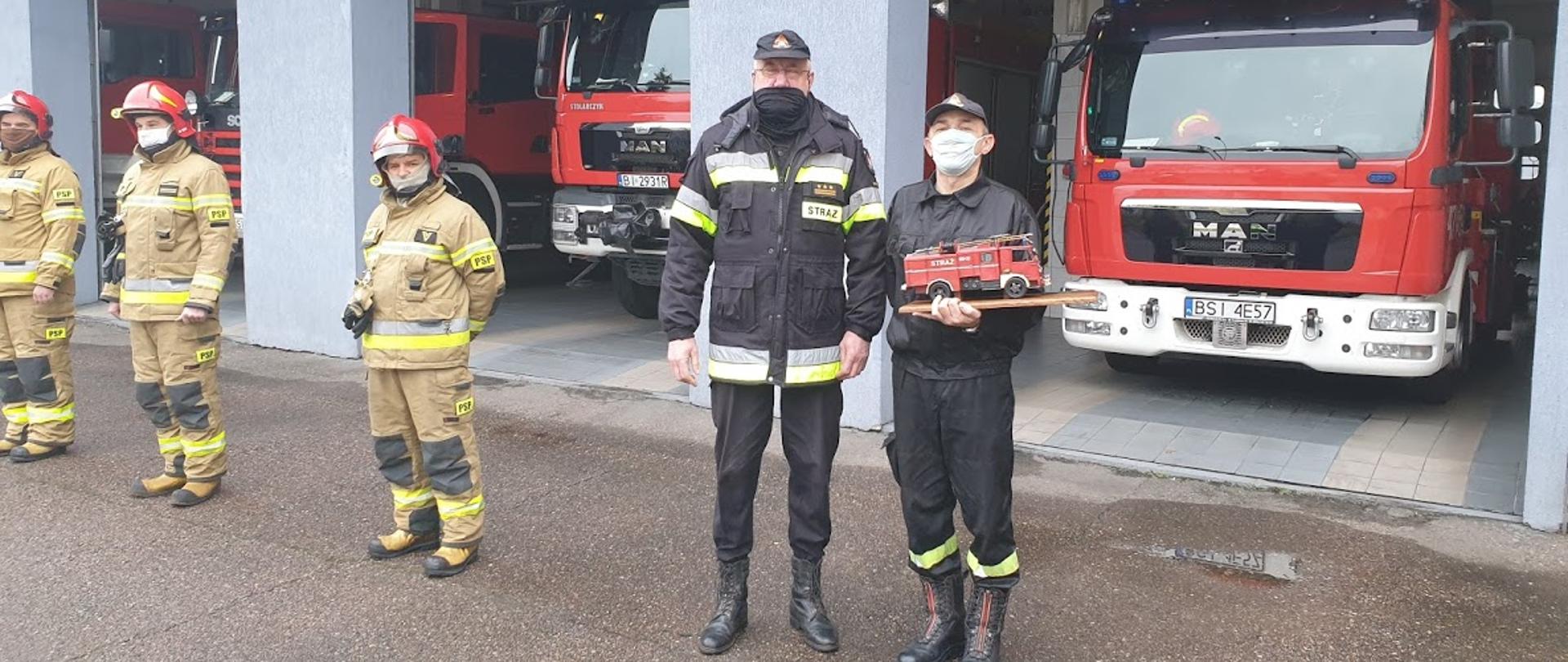 Na zdjęciu strażak w ubraniu koszarowym trzymający pamiątkowy model zabytkowego samochodu strażackiego. Obok niego po lewej stoi Komendant Powiatowy PSP w Siemiatyczach w ubraniu dowódczo-sztabowym. Dalej po lewej trzech strażaków w ubraniach specjalnych. Wszyscy stoją na tle otwartych garaży strażnicy PSP w Siemiatyczach, w garażu widać cztery czerwone samochody ratowniczo-gaśnicze.