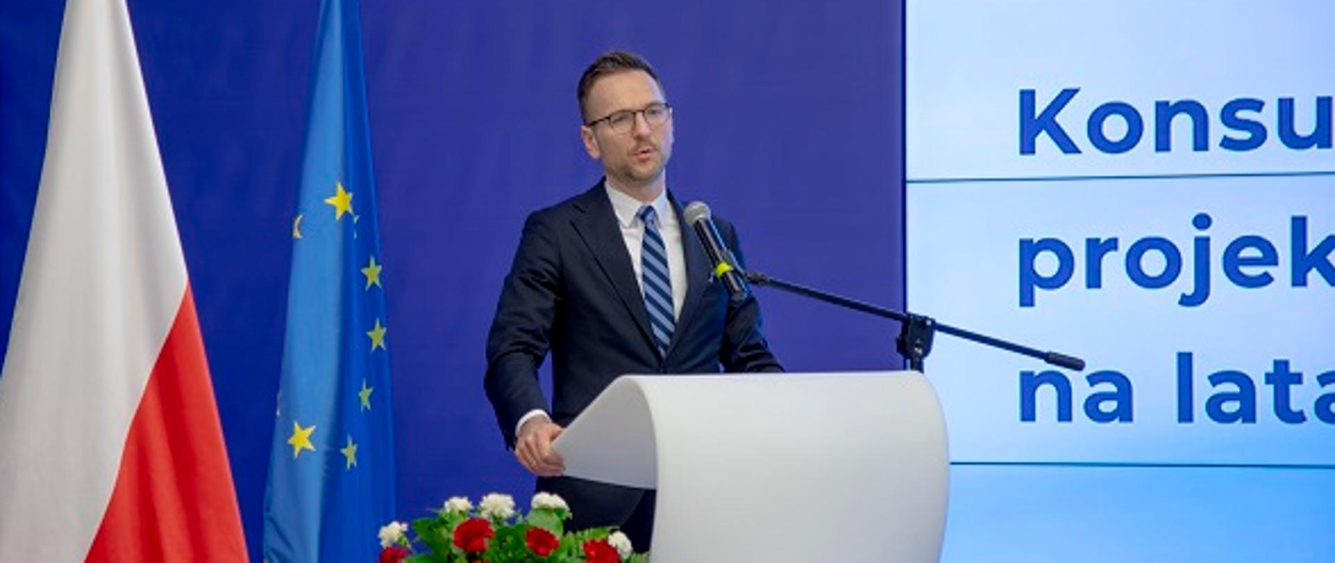 Minister Waldemar Buda przemawia przy mównicy, za nim flagi Polski i UE oraz napis Ministerstwo Funduszy i Polityki Regionalnej