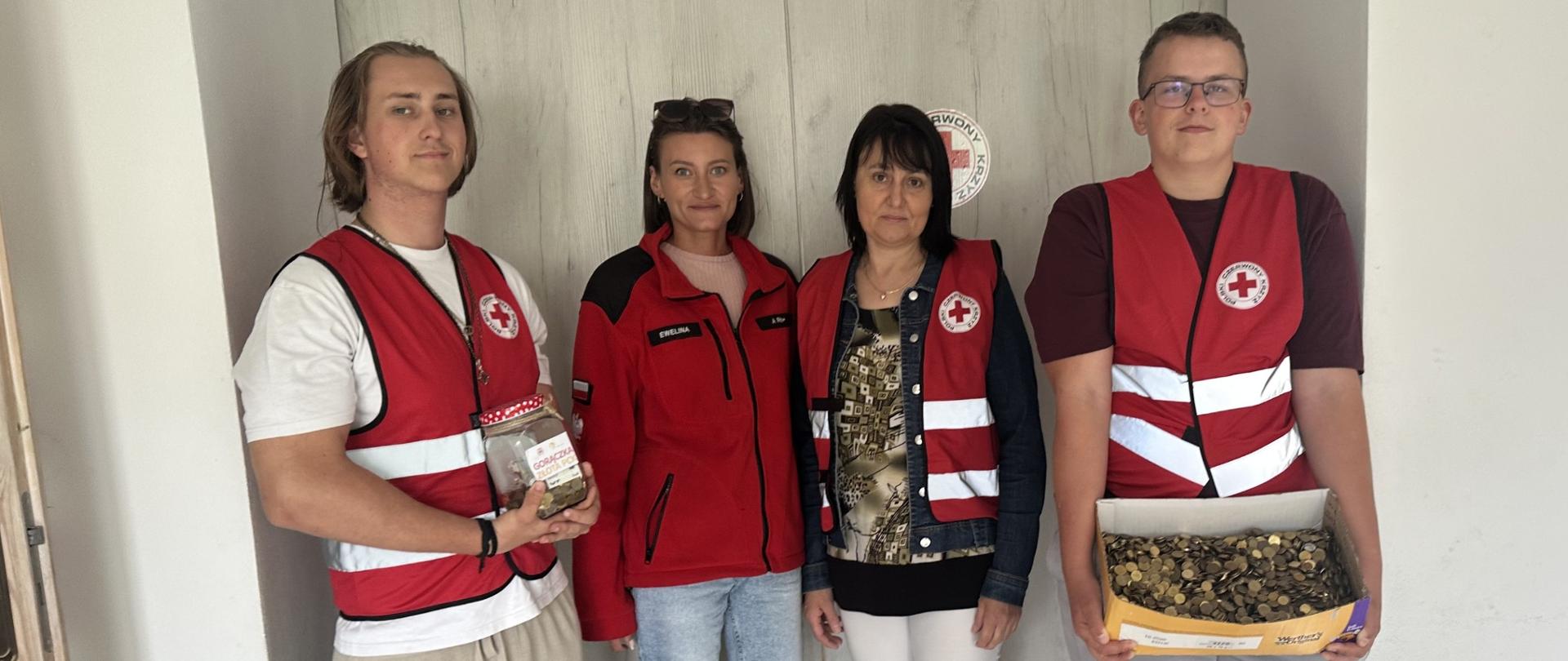 Grupa wolontariuszy PCK w kamizelkach prezentuje zebrane monety w słoiku i pudełku.
