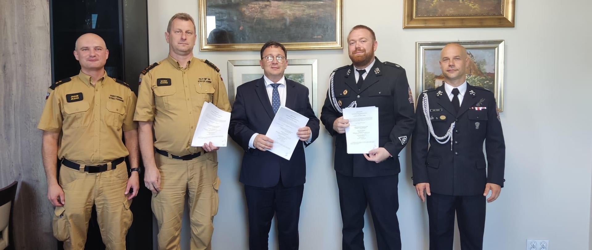 Na zdjęciu pięć osób stojących obok siebie, trzy z nich trzyma w dłoniach przed sobą dokumenty