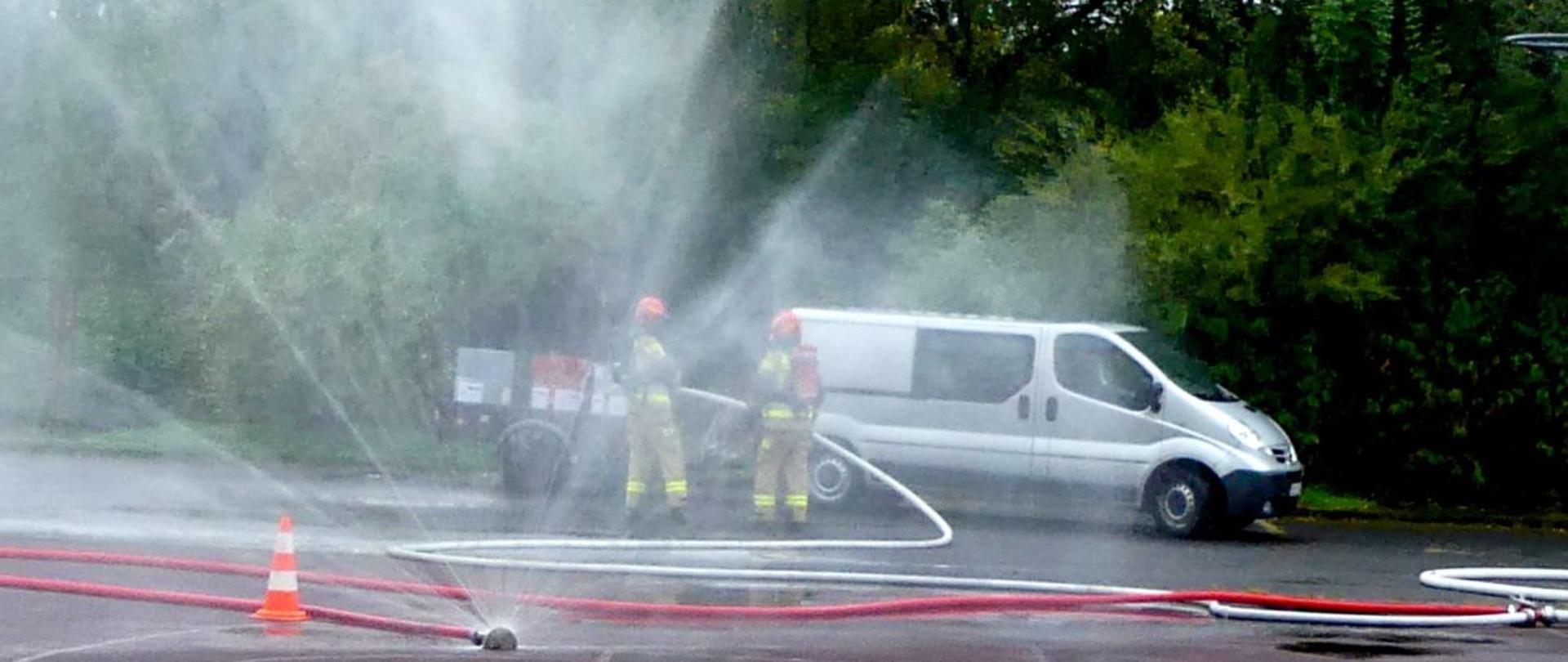 Na zdjęciu widać dwóch strażaków w żółtych mundurach i czerwonych kaskach, którzy stoją obok siebie. Znajdują się na zewnątrz, w pobliżu białego busa zaparkowanego na tle zielonych drzew. Na pierwszym planie widoczne są rozwinięte węże strażackie, z których tryska woda pod dużym ciśnieniem, tworząc rozpryski i mgłę wodną w powietrzu. Na ziemi stoi także pomarańczowy pachołek ostrzegawczy.