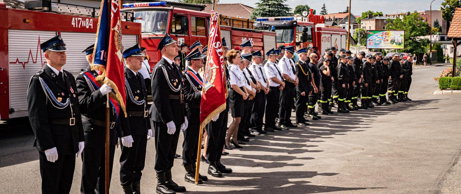 Na pierwszym tle zdjęcia widoczni są strażacy - druhowie Ochotniczej Straży Pożarnej, wśród nich widać dwa poczty sztandarowe. Strażaków ubranych w mundury wyjściowe oraz koszarówki.
Pozują na tle samochodów ratowniczo-gaśniczych. 