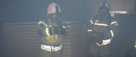 Pożar w tartaku. Wnętrze jednego z budynków produkcyjnych, w którym znajdują się sztaple tarcicy. Przed nimi dwóch strażaków w ubraniach koloru piaskowego i w białych hełmach z czerwonymi elementami, którzy prowadzą działania ratowniczo-gaśnicze. mają na sobie założony sprzęt ochrony układu oddechowego. Jeden z nich trzyma w rękach prądownicę podłączoną do węża. . Do ubrania ma przypiętą żółtą latarkę.