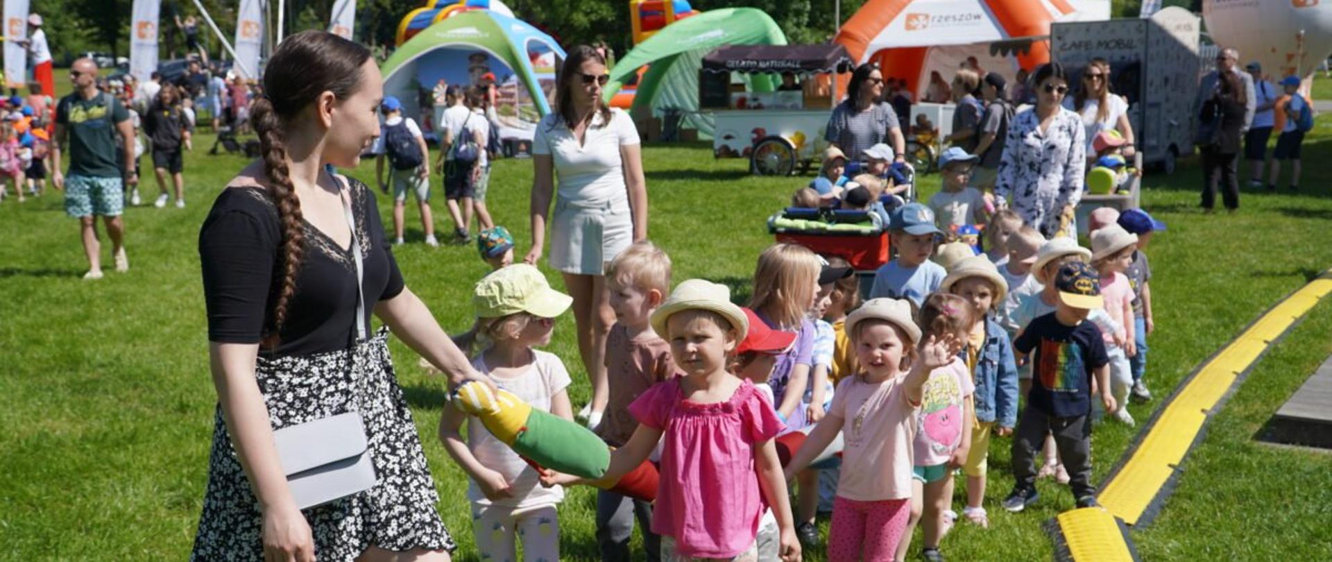 Młoda kobieta prowadząca dzieci przedszkolne podczas imprezy plenerowej z okazji Dnia Dziecka. W tle stoiska edukacyjne