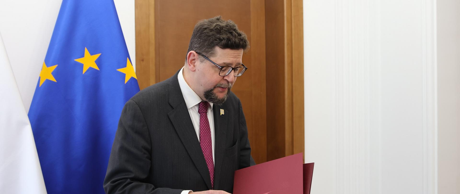 Wiceminister Szeptycki stoi i trzyma czerwoną teczkę.