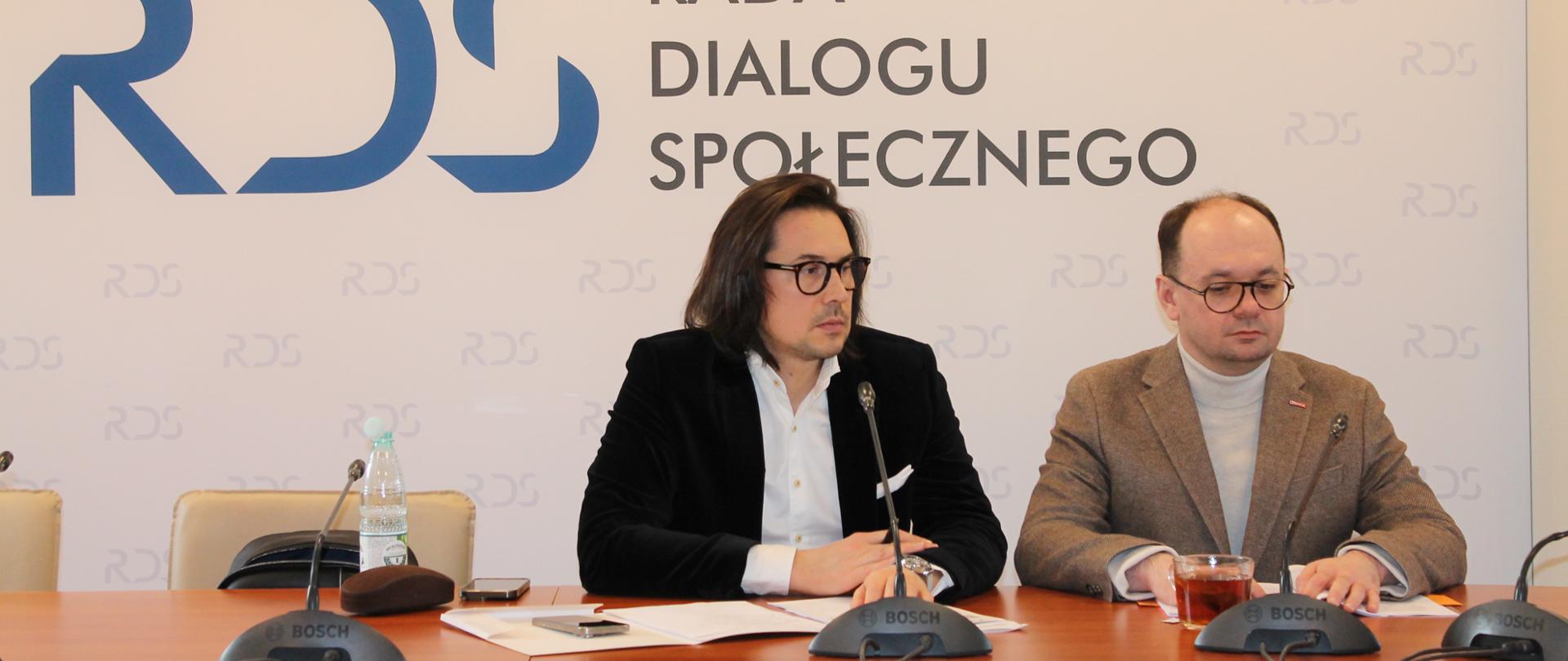 Na zdjęciu jest stół prezydialny, za którym od lewej strony siedzi Przewodniczący Zespołu Błażej Mądrzycki oraz Sebastian Gajewski – Podsekretarz Stanu w Ministerstwie Rodziny, Pracy i Polityki Społecznej. W tle siedzących widnieje baner z napisem Rada Dialogu Społecznego. 