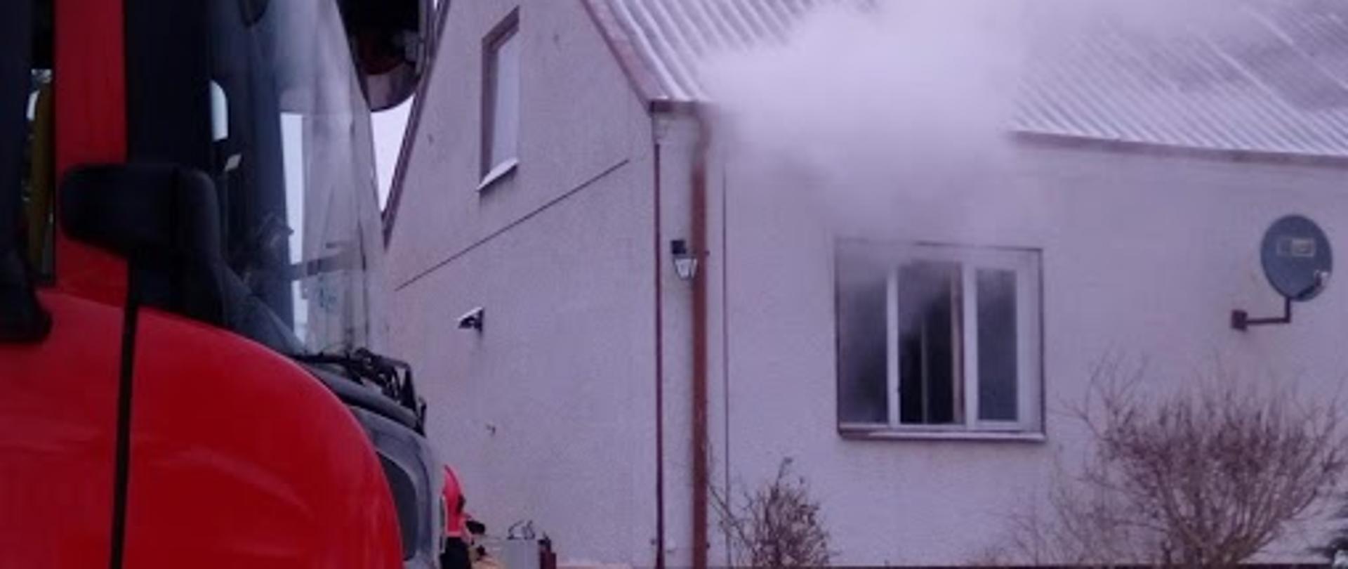 Zdjęcie przedstawia przód kabiny samochodu gaśniczego stojącego na zaśnieżonej drodze przed wjazdem na posesję, na drugim planie biały budynek mieszkalny z czerwonym dachem z widocznymi kłębami białego dymu wydobywającego się z otwartych okien jednego z pomieszczeń budynku.