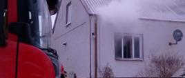 Zdjęcie przedstawia przód kabiny samochodu gaśniczego stojącego na zaśnieżonej drodze przed wjazdem na posesję, na drugim planie biały budynek mieszkalny z czerwonym dachem z widocznymi kłębami białego dymu wydobywającego się z otwartych okien jednego z pomieszczeń budynku.