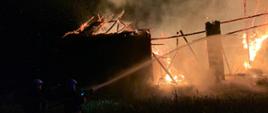 Widoczne miejsce zdarzenia płonący budynek stodoły ze zniszczoną konstrukcją dachu widać dym i płomienie oraz strażacy prowadzący działania gaśnicze.