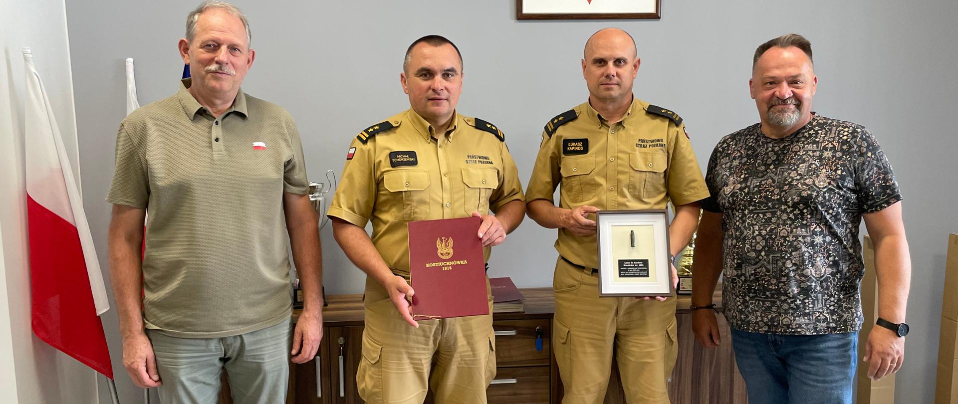 Czterech mężczyzn stoi w gabinecie Komendy Powiatowej PSP w Mielcu. Dwóch strażaków w umundurowaniu służbowym stoi w środku – jeden trzyma teczkę z napisem Kostiuchnówka 1916, drugi ramkę z pamiątkową łuską. Po bokach stoją dwaj mężczyźni w ubraniach cywilnych. W tle widoczny herb Polski.