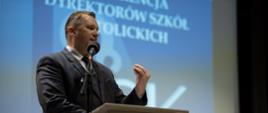 Minister Czarnek stoi przy mównicy i mówi do mikrofonu, za nim wielki ekran z białym napisem Konferencja Dyrektorów Szkół Katolickich.