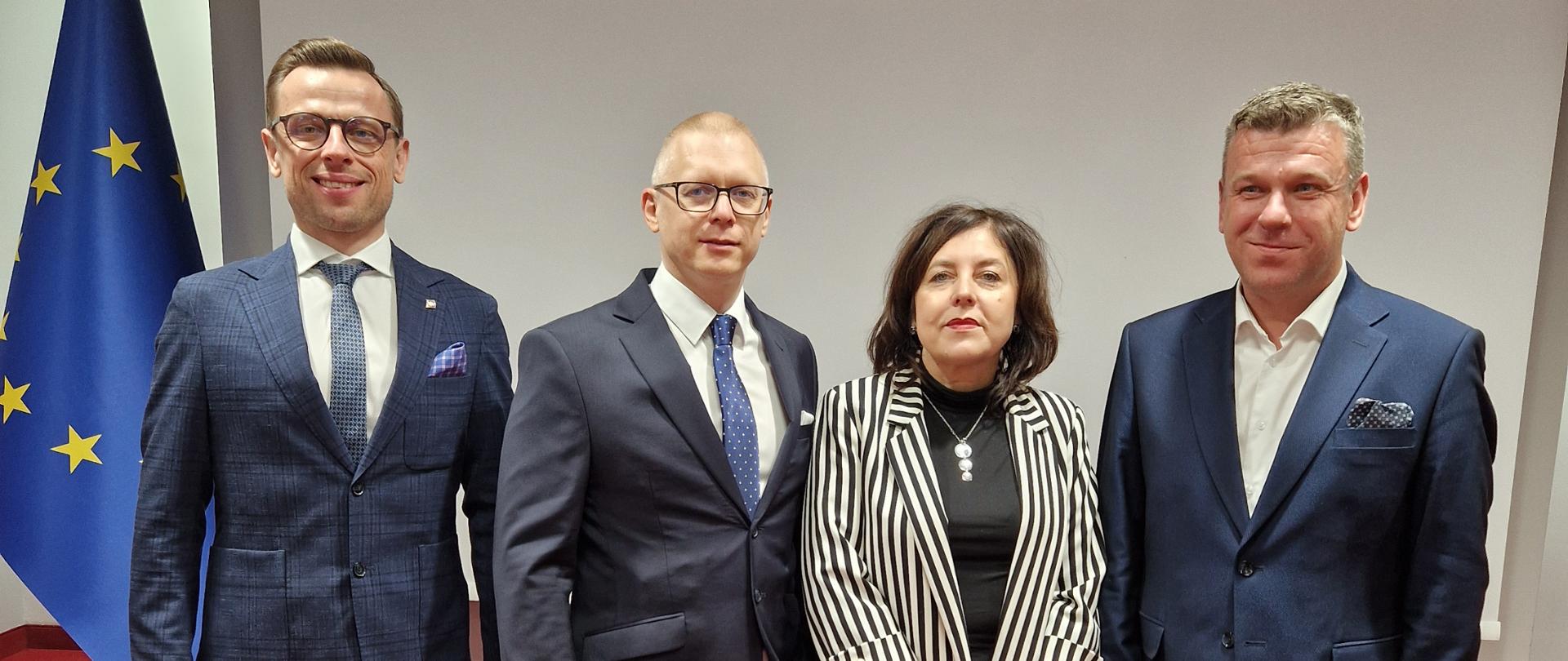 Grupa czterech osób pozuje do formalnego zdjęcia na tle flagi Unii Europejskiej. Dwóch mężczyzn stojących po lewej stronie ma na sobie ciemne garnitury i niebieskie krawaty, obaj w jasnych koszulach. Między nimi stoi kobieta w czarno-białym, pasiastym stroju, a obok niej znajduje się mężczyzna w niebieskim garniturze.