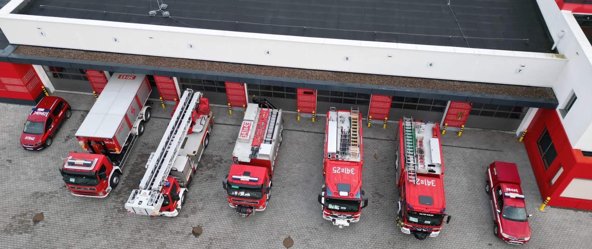 Widoczny z lotu ptaka plac wyjazdowy przed jednostką ratowniczo- gaśniczą numer 1 w Elblągu. Wszystkie bramy garażowe są otwarte, a przed nimi stoją pojazdy pożarnicze.