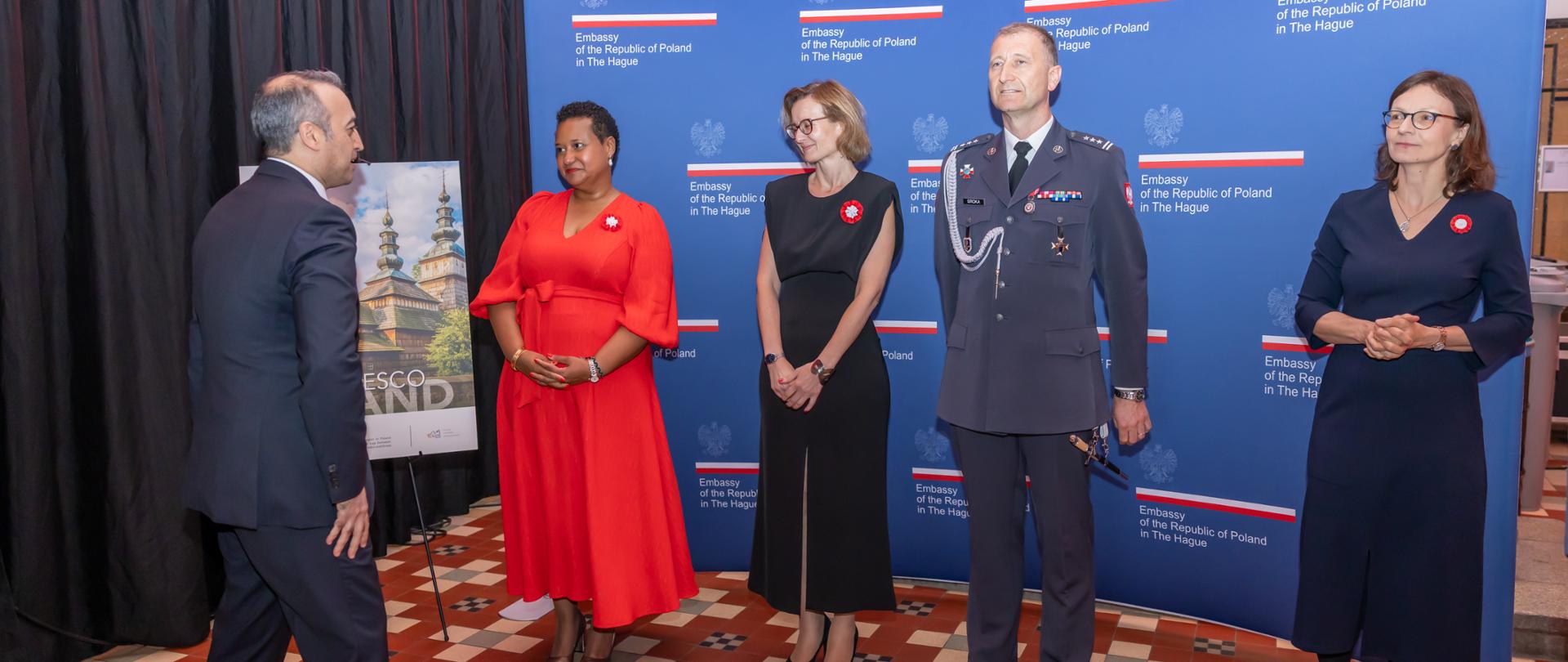 Ambasador Margareta Kassangana, w czerwonej sukience, wita gości na tle granatowej ścianki z logo ambasady. 