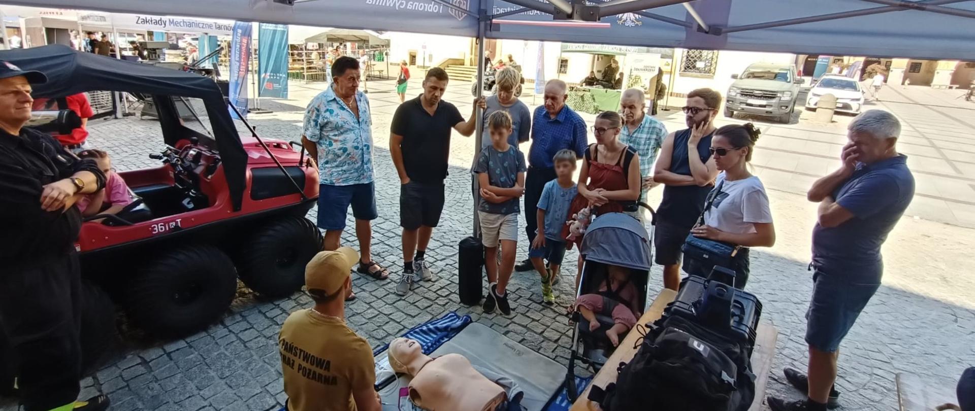 Szerokie ujęcie wnętrza namiotu promocyjnego Państwowej Straży Pożarnej i OLiOC na brukowanym rynku. Grupa przechodniów, w tym dzieci i dorośli, przysłuchuje się pokazowi pierwszej pomocy prowadzonemu przez strażaka klęczącego przy fantomach. Obok namiotu zaparkowany jest czerwony pojazd wielozadaniowy.