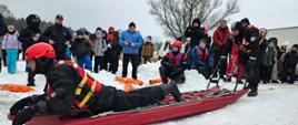 Ratownicy prezentują techniki transportu na saniach lodowych przed grupą dzieci, młodzieży i funkcjonariuszy Policji. Pokaz ma na celu edukację uczestników w zakresie bezpiecznych zachowań i metod ratownictwa zimowego.