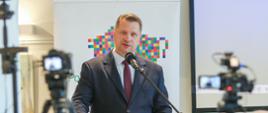 Minister Czarnek stoi i mówi do mikrofonu na stojaku, przed nim zarysy osób siedzących na sali, obok niego dwie kamery na statywach, za nim baner w kolorowy geometryczny wzorek na białym tle.