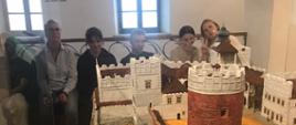 Z przodu duża makieta Zamku Lubelskiego, z tyłu na tle białej ściany z dwoma oknami siedzi sześć osób. 