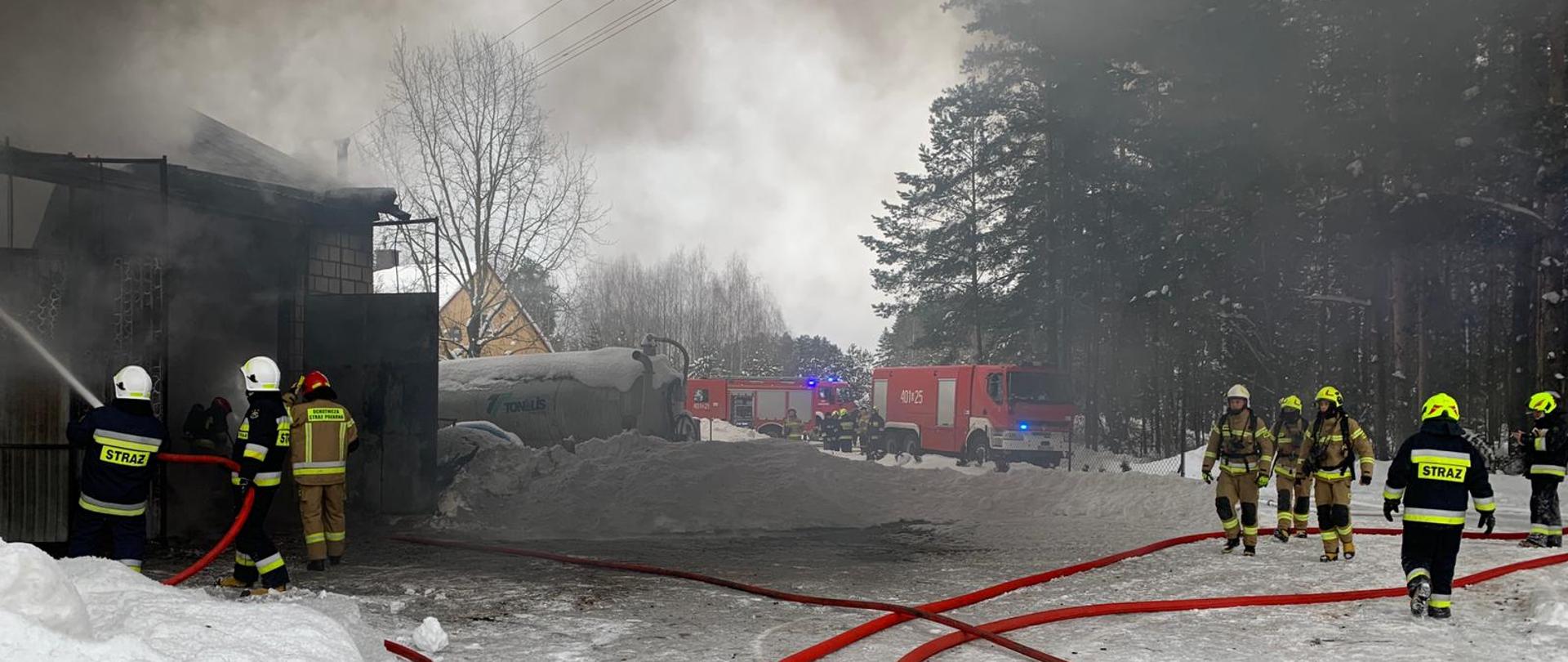 Na zdjęciu widać w tle samochody strażackie; strażacy podają prądy wody do wnętrza budynku