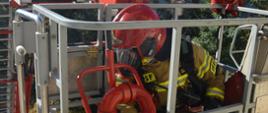 Ćwiczenia w szpitalu. Na pierwszym planie kosz podnośnika hydraulicznego, w którym znajduje się jeden ze strażaków w czerwonym hełmie i piaskowym ubraniu. Kosz jest przy oknie na drugim piętrze. 