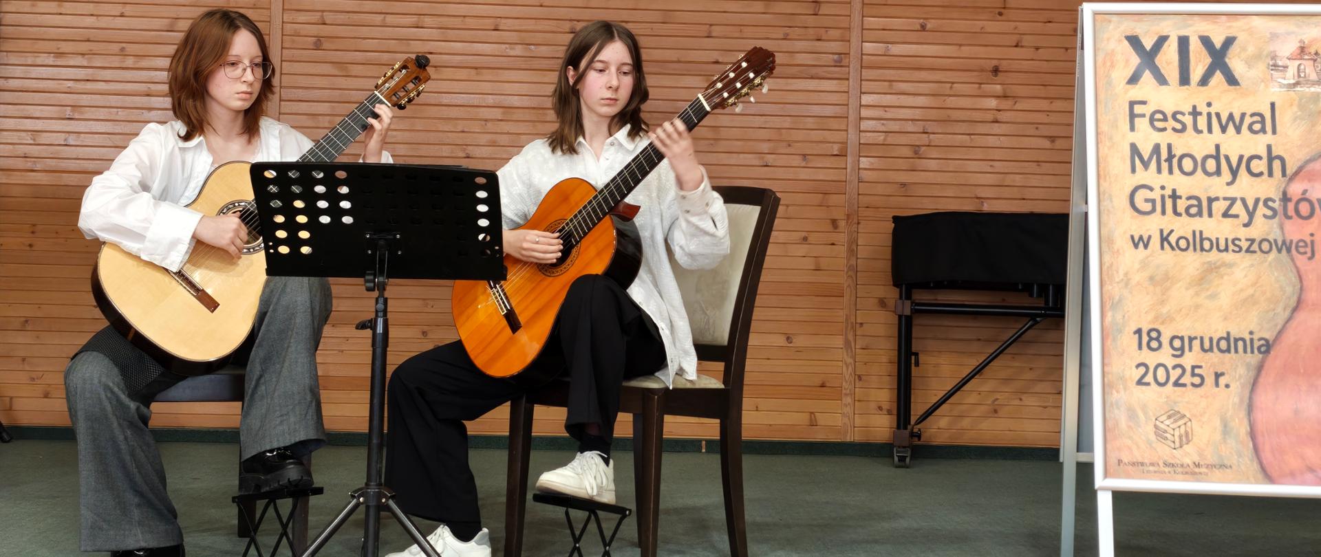 Dwie siedzące na krzesłach dziewczynki grające na gitarach , nogi trzymają na podnóżkach. Przed nimi stoi czarny pulpit, za nimi ściana w kolorze brązowym z godłem państwowym i napisem w Kolbuszowej. Z prawej strony widać fragment baneru reklamującego 19 Festiwal Młodych Gitarzystów w Kolbuszowej