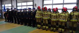 Zdjęcie wykonano za dnia w garażu Komendy Powiatowej Państwowej Straży Pożarnej w Sępólnie Krajeńskim, Na tle bram wjazdowych od lewej stoi poczet sztandarowy, dalej w dwuszeregu strażacy ubrani w mundury wyjściowe a dalej strażacy ubrani w ubrania specjalne i hełmy. 