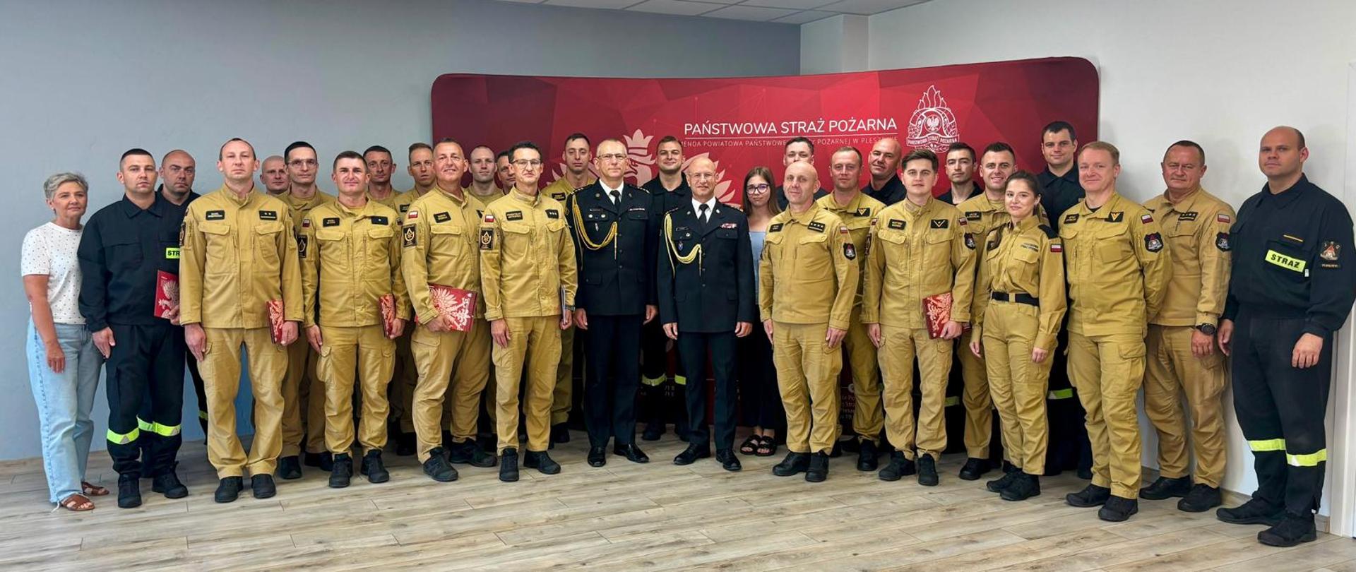 Szerokie zdjęcie grupowe kilkudziesięciu strażaków oraz pracowników cywilnych zgromadzonych w sali konferencyjnej na tle czerwonej ścianki PSP. Większość strażaków ubrana jest w piaskowe mundury służbowe oraz czarne mundury koszarowe i wyjściowe. Wszyscy patrzą w stronę aparatu.
