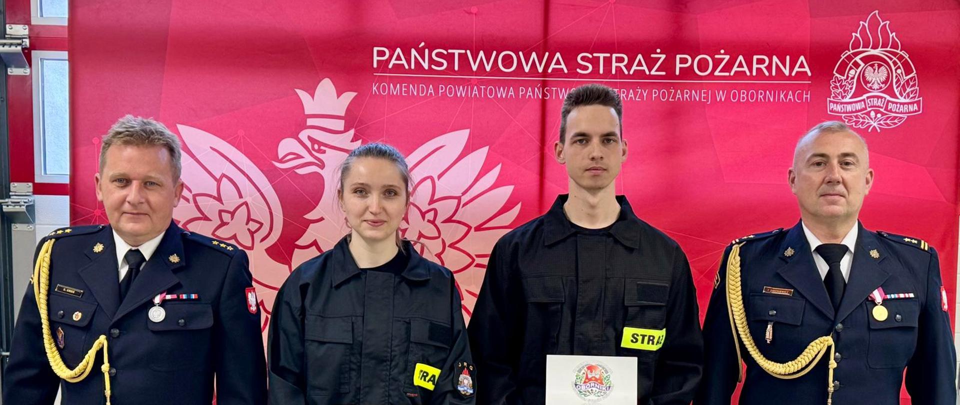 Cztery osoby pozują do wspólnego zdjęcia na tle czerwonego baneru Komendy Powiatowej Państwowej Straży Pożarnej w Obornikach. Pośrodku stoją dwie osoby w czarnych ubraniach strażackich, trzymające pamiątkowe publikacje z wizerunkiem komendy. Po bokach znajdują się dwaj funkcjonariusze PSP w galowych mundurach.