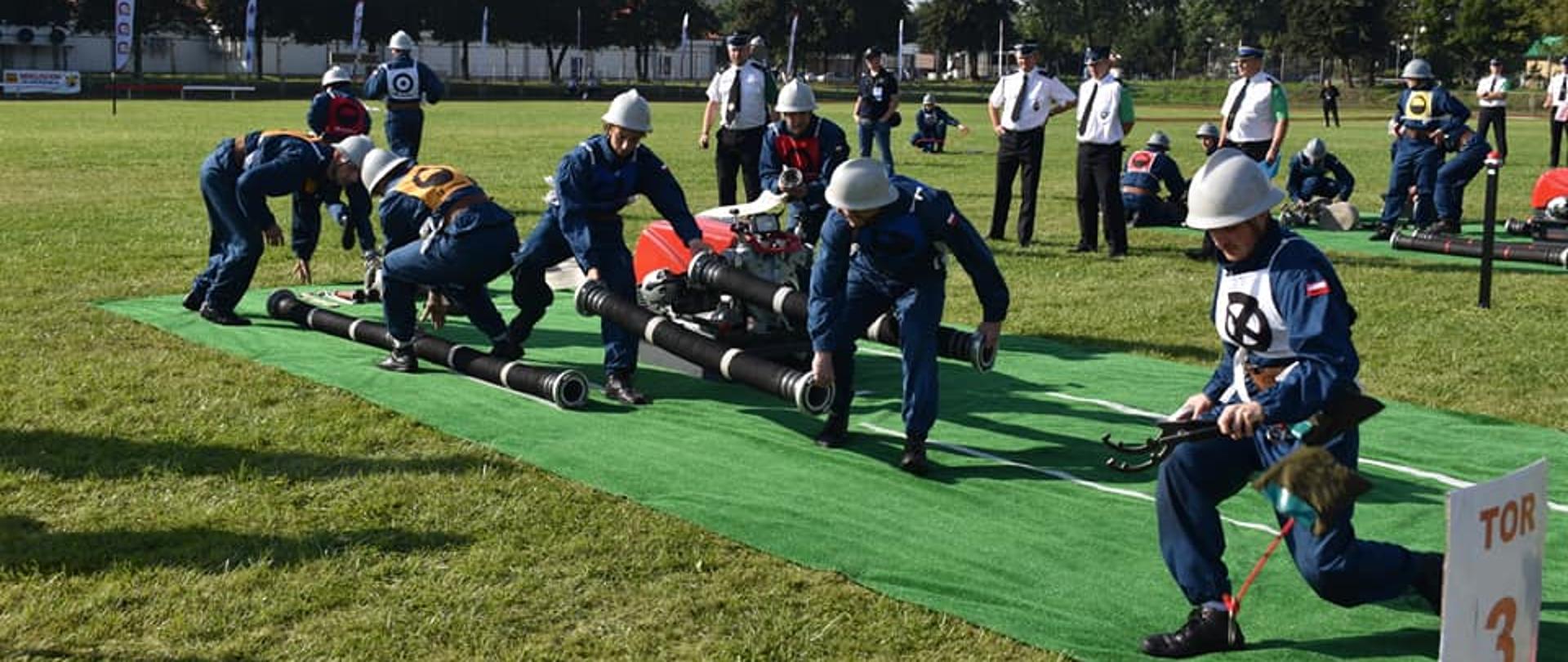 Zdjęcie przedstawia zawodników OSP podczas startu w zawodach sportowo-pożarniczych, które odbyły się w dniach 27-28 sierpnia 2021 roku w Koninie. 