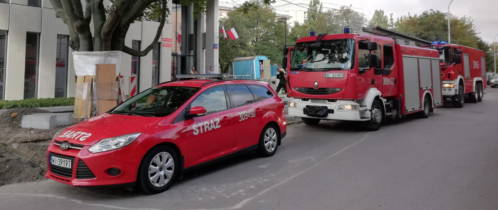 Na zdjęciu widoczne są pojazdy straży pożarnej w tle drzewo oraz budynek biurowy.