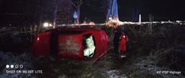 Zdjęcie przedstawia miejsce wypadku czerwonego samochodu osobowego w porze nocnej. Samochód leży na lewym boku, poza drogą. Obok samochodu stoi mężczyzna ubrany na czarno oraz ratownik medyczny ubrany na czerwono.