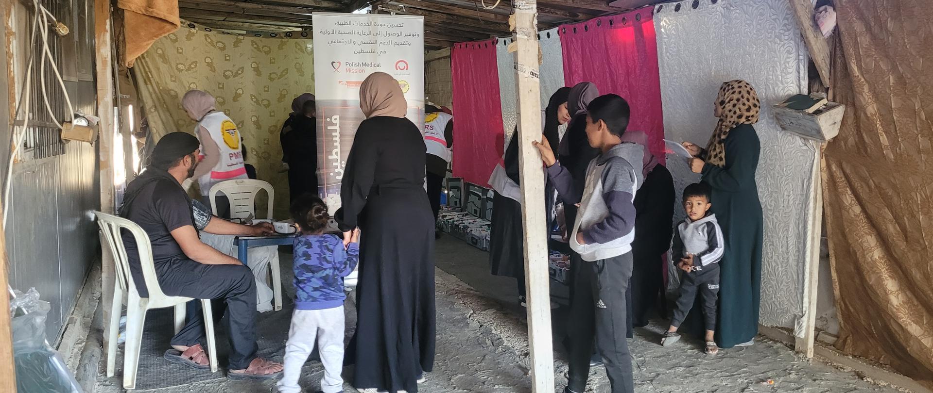W dużym namiocie kobiety, dzieci i mężczyźni korzystają z pomocy lekarskiej. Widać personel medyczny w kamizelkach z napisem PMM. Mężczyzna ma badane ciśnienie krwi