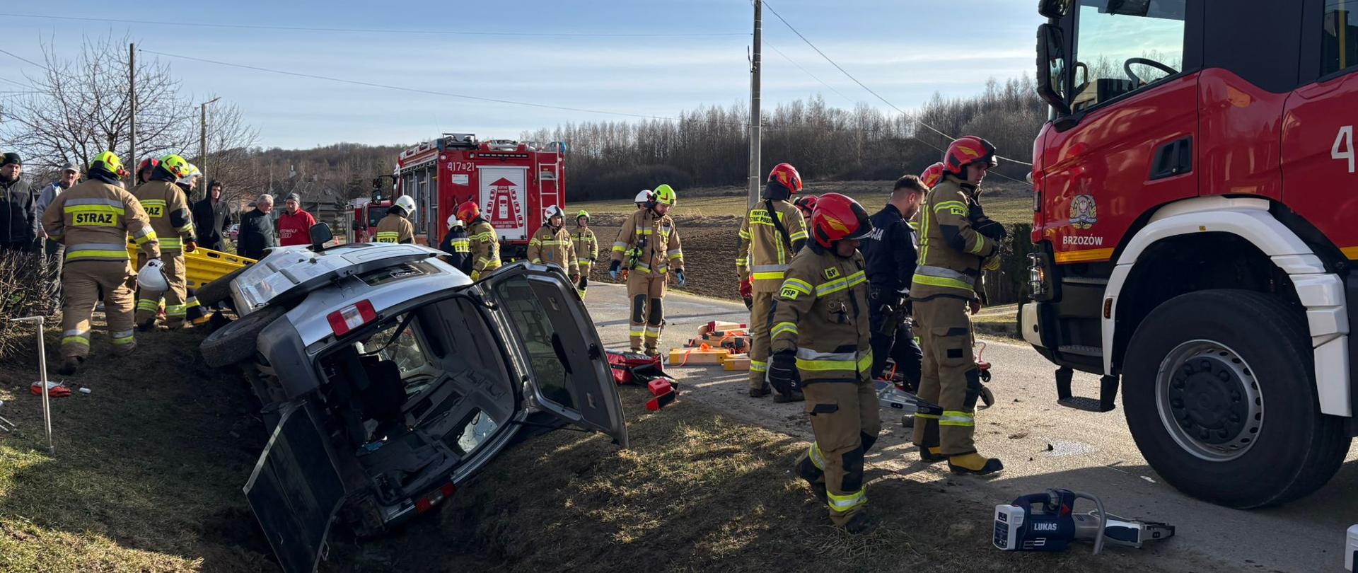 Kolorowa fotografia wykonana w pogodny dzień na drodze powiatowej. Przedstawia samochód osobowy w rowie na prawym boku z otwartym bagażnikiem, obok pojazdu znajduje się część samochodu pożarniczego oraz strażacy w ubraniach specjalnych i hełmach.
