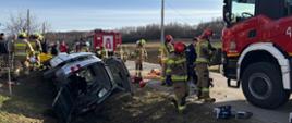 Kolorowa fotografia wykonana w pogodny dzień na drodze powiatowej. Przedstawia samochód osobowy w rowie na prawym boku z otwartym bagażnikiem, obok pojazdu znajduje się część samochodu pożarniczego oraz strażacy w ubraniach specjalnych i hełmach.
