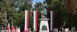 Rocznica wybuchu II WŚ Zdjęcie przedstawia plac, na którym stoi pomnik Żołnierza Polskiego. Pomnik ustawiony jest na tle drzew na cokole, na którym umieszczone jest godło RP. Pomnik to postać żołnierza w plaszczu i czapce rogatywce, trzymającego karabin. Za pomnikiem na pionowych masztach zawieszone są 4 flagi biało-czerwone. Z lewej strony w szeregu stoją poczty sztandarowe. Obok pomnika znajdują się wartownicy.