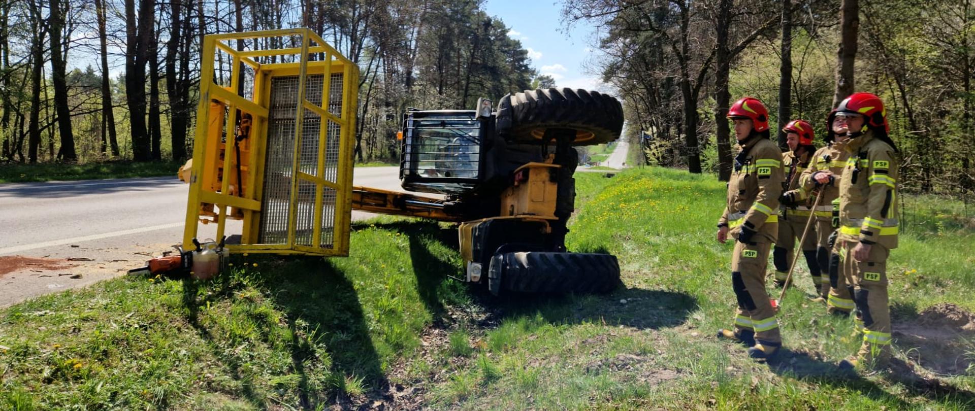 Zdjęcie wykonane na drodze wojewódzkiej numer 740 w miejscowości Kosów. Na zdjęciu widać przewrócony na bok w przydrożnym rowie podnośnik koszowy. Maszyna w kolorze żółtym. Po obu stronach drogi znajduje się zielony las. Po prawej stronie od podnośnika na poboczu stoi czterech strażaków. Ubrani są w czerwone hełmy oraz ubrania specjalne w kolorze żółtym piaskowym. 